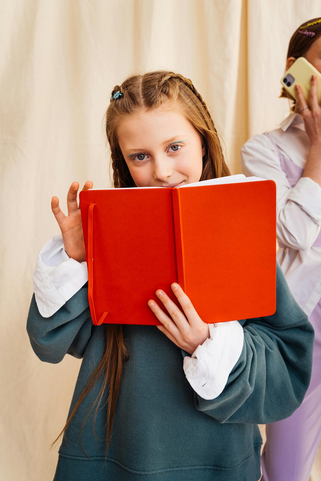 Girl peeking over an orange notebook while her friend talks on a phone behind her